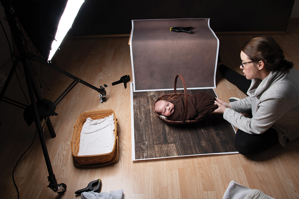 Photographer sitting on the floor next to a baby in a basket in preparation for a photo session.