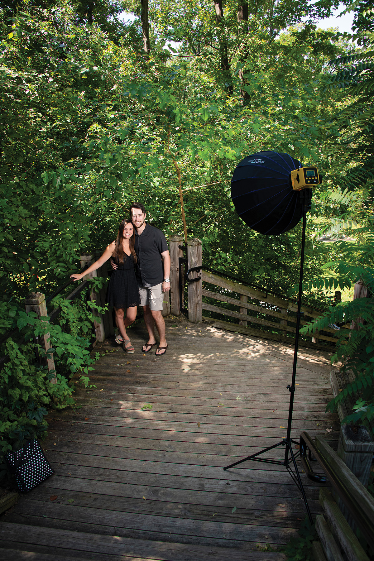 A couple poses for a portrait on a boardwalk in a wooded area. A flash and umbrella on a light stand are set up in front of them.