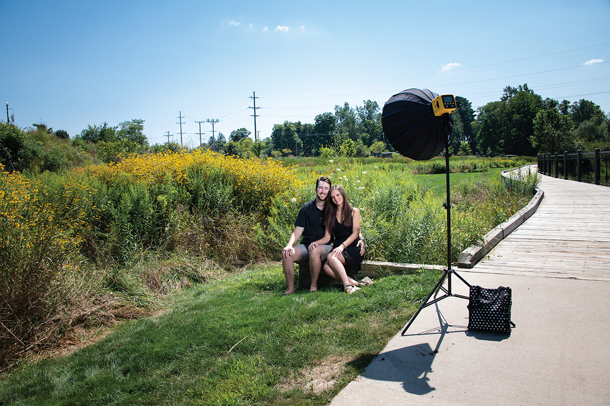 A couple sitting trailside in front of flowers with a light stand and flash with umbrella set up in front of them