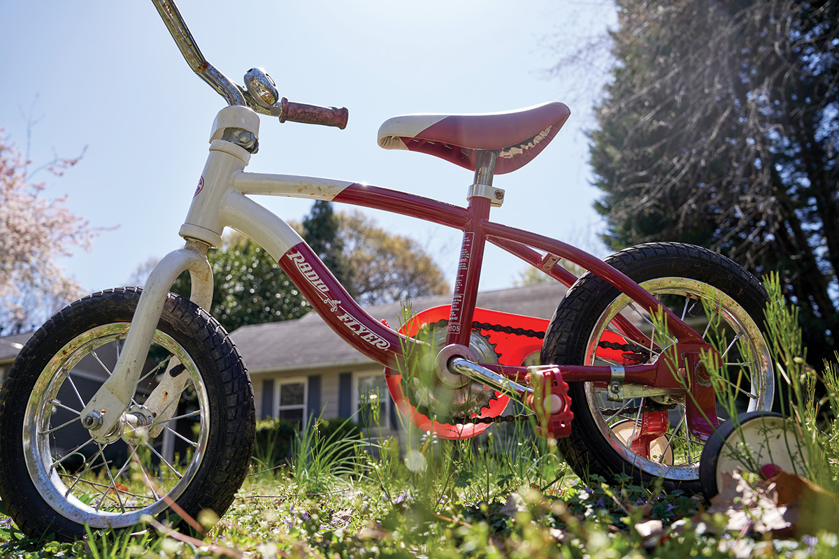 Red and white child's bicycle in a grassy front yard, viewed from an angle low to the ground, and with a house and trees in the background. The sun is backlighting the bike.