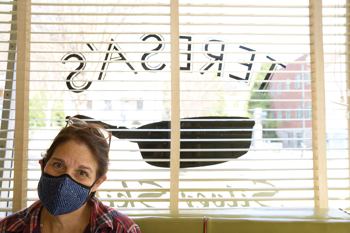 A white woman with brown hair pulled back and glasses on top of her head, wearing a mask, sitting inside a restaurant in front of the front window with open blinds. The name of the restaurant is painted on the window, seen in reverse 