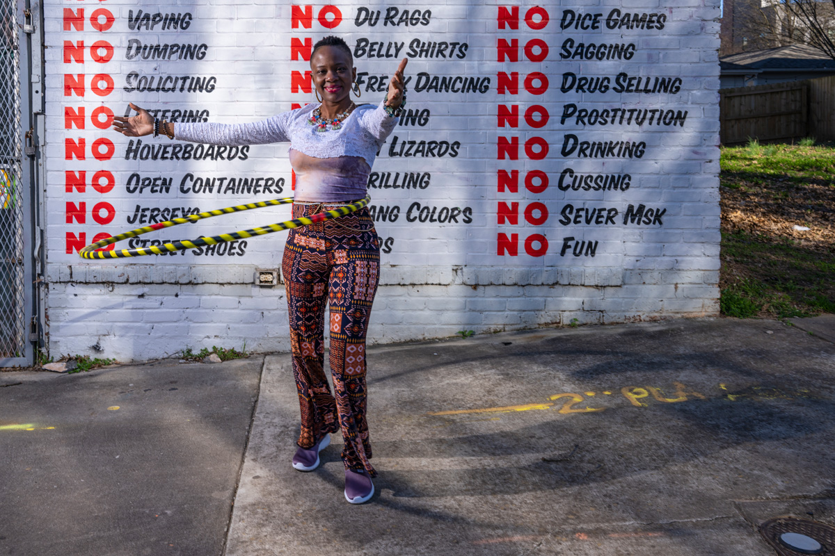 A Black woman with short hair in colorful print pants and a blue top using a yellow and black Hula Hoop on the sidewalk in front of a blue brick wall with many statements about what is not allowed, each starting with NO in red letters