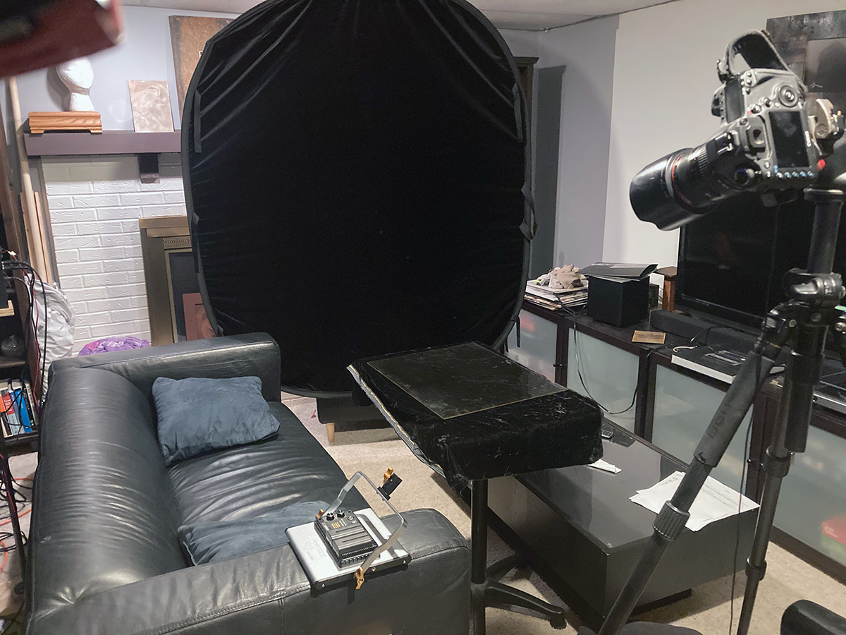 A home living room space with a black velvet backdrop, camera on tripod, and a still life staging platform crowded between a couch, TV, and coffee table. An LED light is face down on one arm of the couch.