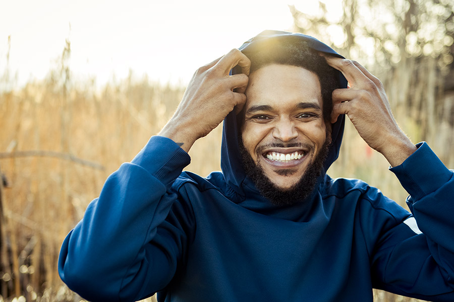 A young person with light brown olive skin tone, mustache and goatee, smiling, wearing a blue hoodie sweatshirt. There is golden tall grass in the background. The subject has both hands on the hoodie as if in the act of pulling it back. 