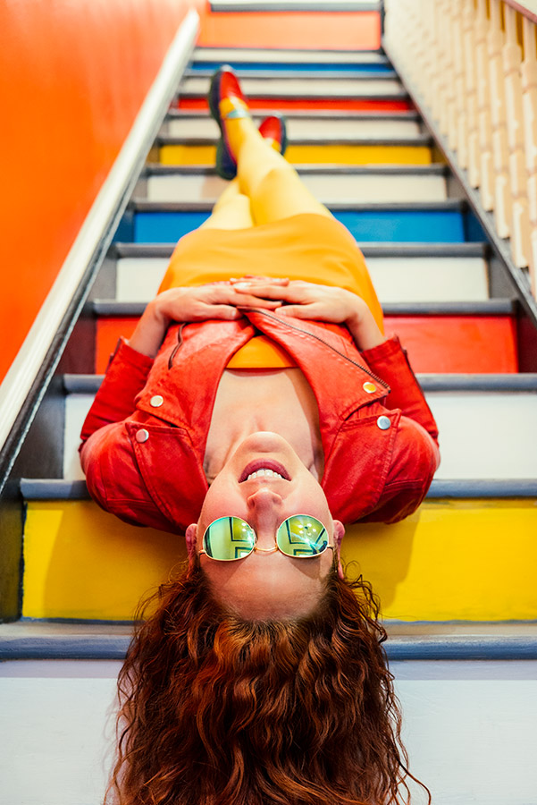 A person with red hair, sunglasses, orange jacket, and a yellow skirt lying on brightly colored steps with their head in the foreground and feet at the further perspective. Their head is upside down in the viewer's POV.