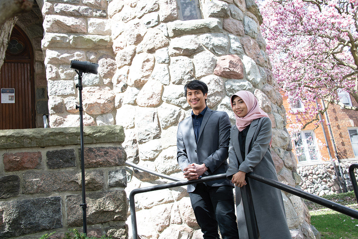 A couple stands in front of a light stand with StellaPro light mounted on it. The location is outdoors in front of a stone building with blossoming trees in the background.