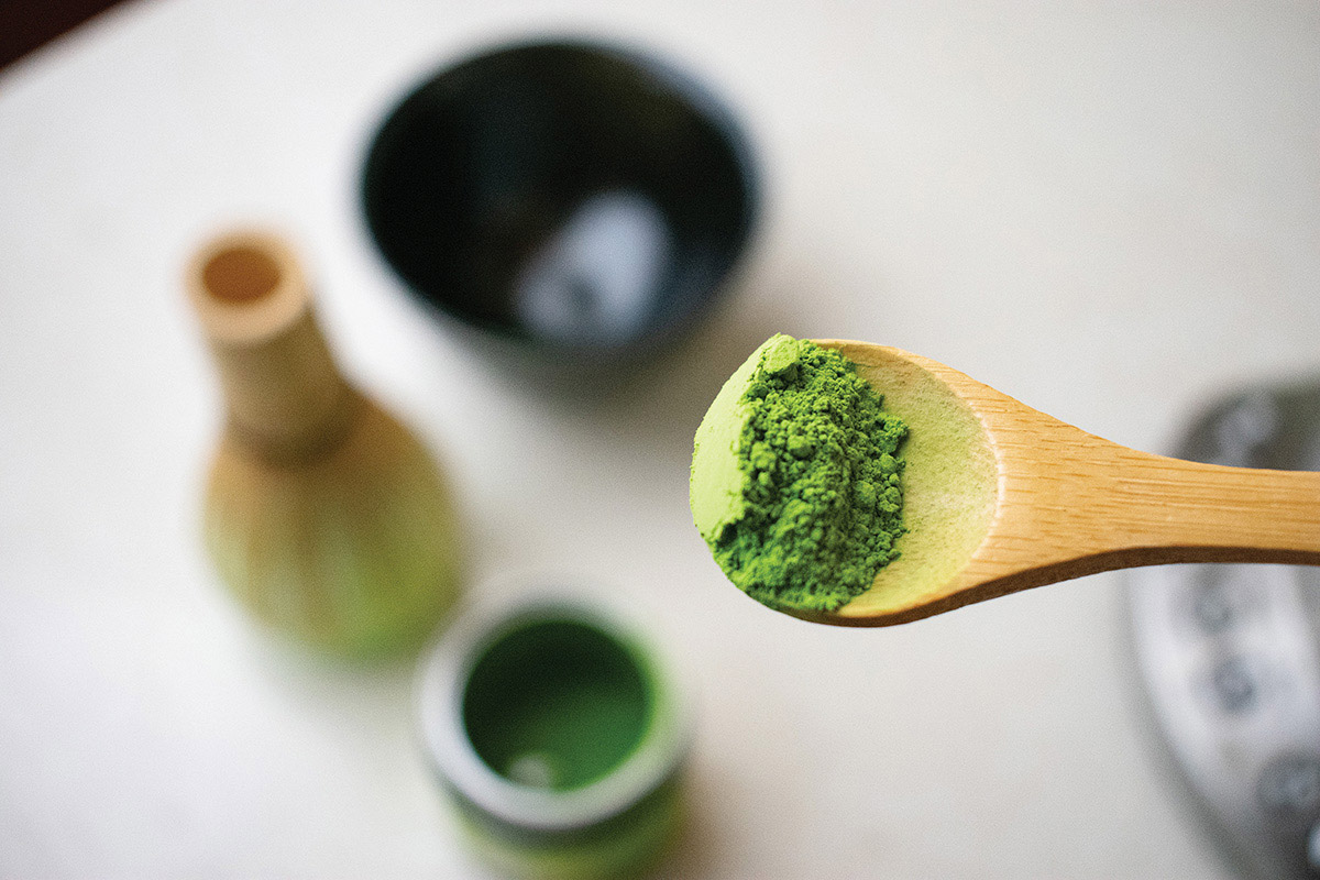 Matcha tea on a wooden spoon with other tea related items in the background, out of focus
