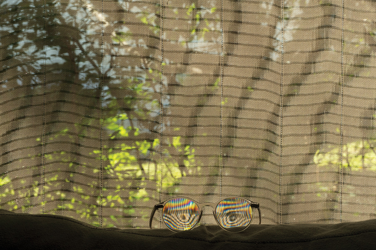Woven window blinds backlit with dappled, shaded sunlight and a pair of eyeglasses sitting in the foreground with colorful patterns reflected in the lenses.