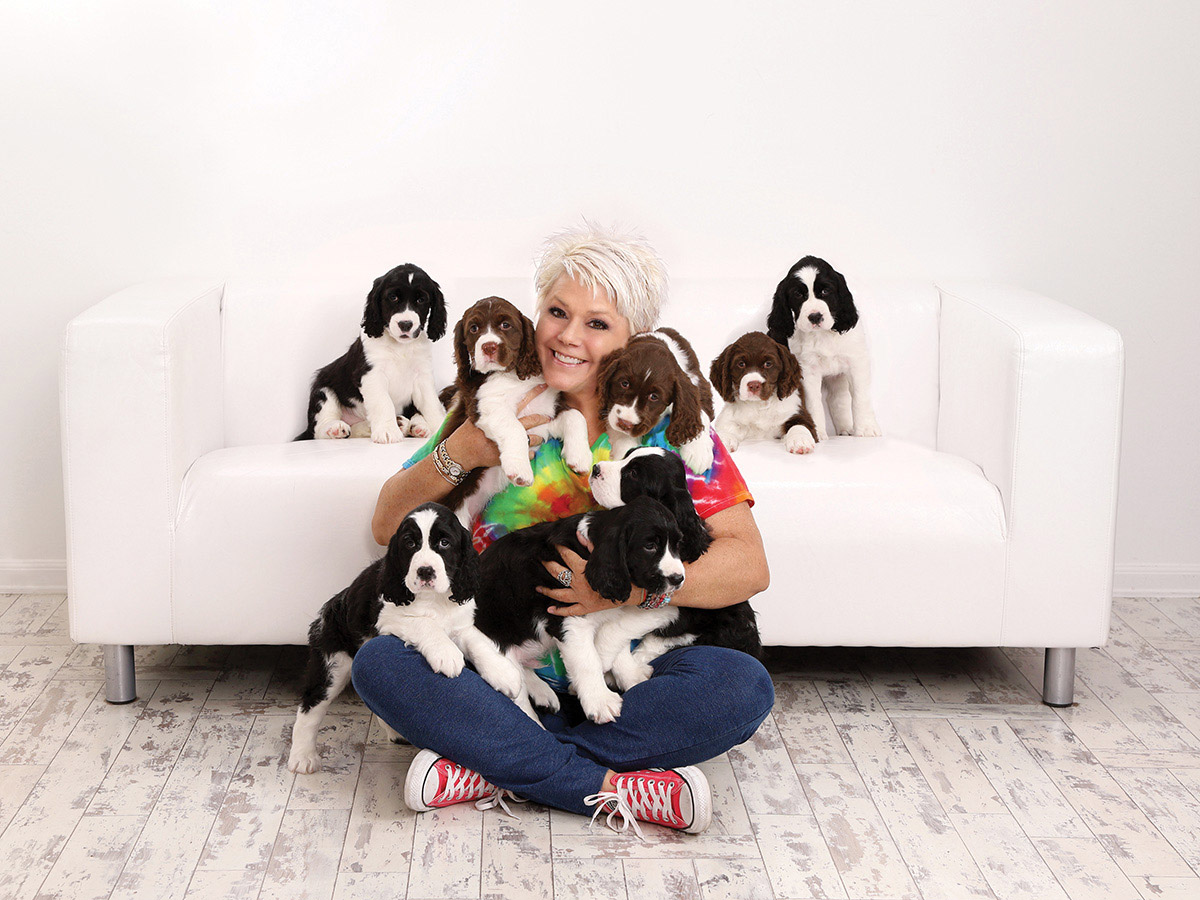 Nine puppies surround photographer Kay Eskridge as she sits on the floor with her back against a white couch. 