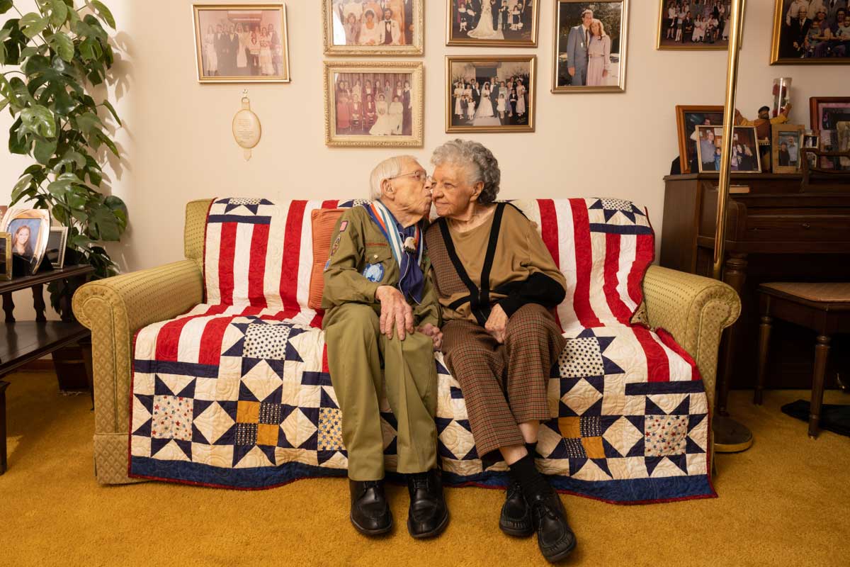A man and woman couple, identified as centenarians, sit on the couch in their living room. He is kissing her cheek, and there are family photos on the wall behind them. 