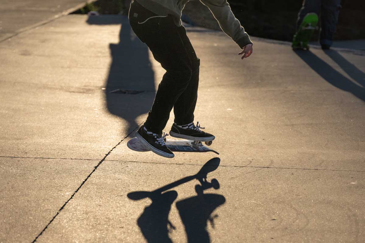 Late day sun backlights a skateboarder doing a board flip, shown from waist down