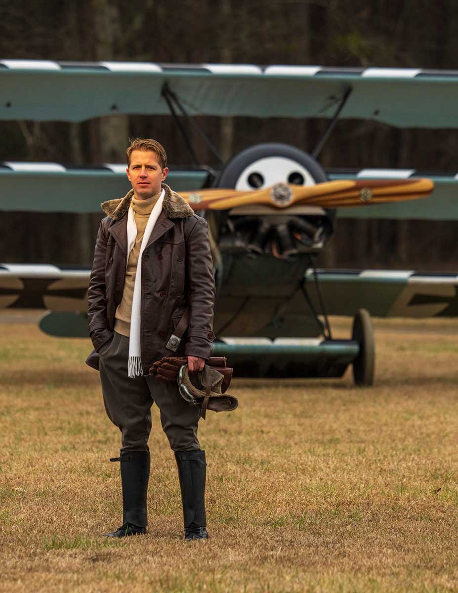 A man dressed in World War I period pilot costume stands in a field in front of a vintage triplane.