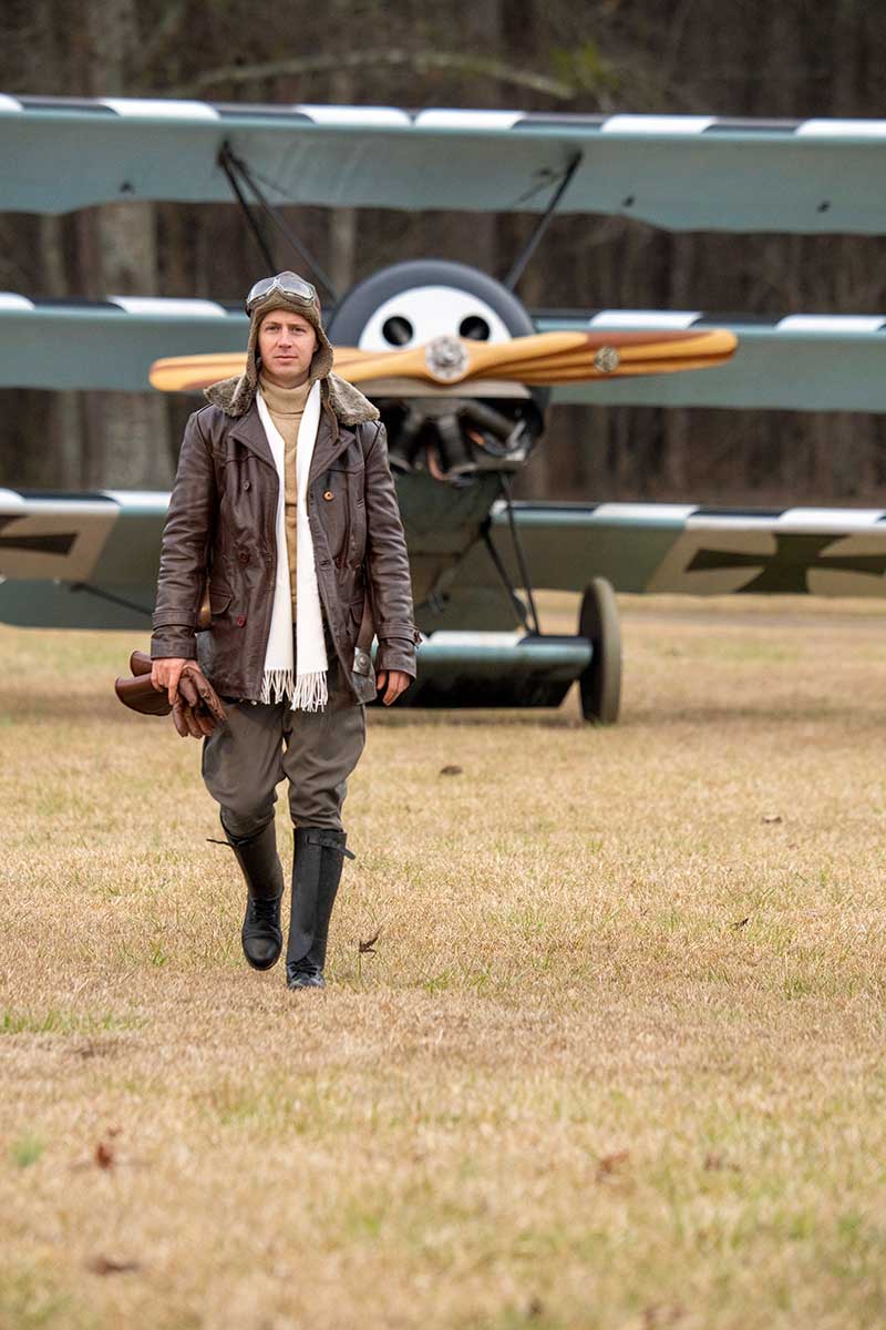 Man dressed as WWI era pilot walks toward viewer in a field in front of a Fokker triplane