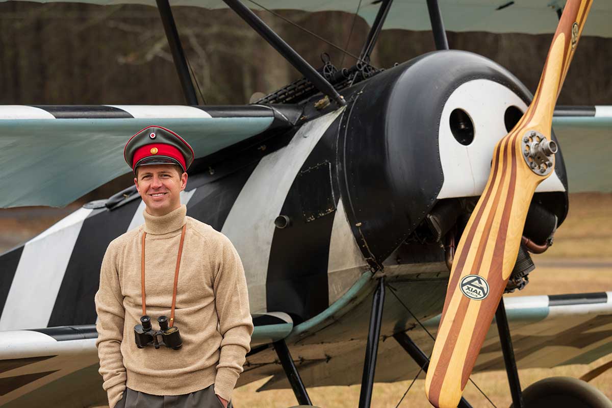 A man with vintage binoculars hung around his neck and wearing tan turtleneck and WWI era military pilot hat stands in front of a restored Fokker Triplane