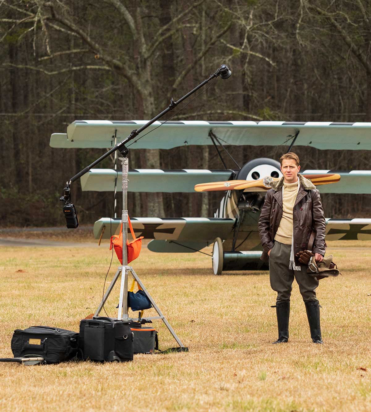 A man dressed in World War I era pilot costume stands in a field in front of a vintage Fokker Triplane. Lighting equipment is shown in the foreground.