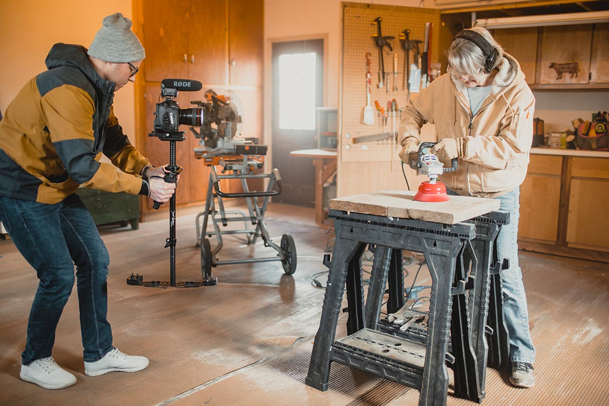A young man with DSLR camera on a stabilizer with microphone mounted videos an older woman sanding wood on sawhorses.