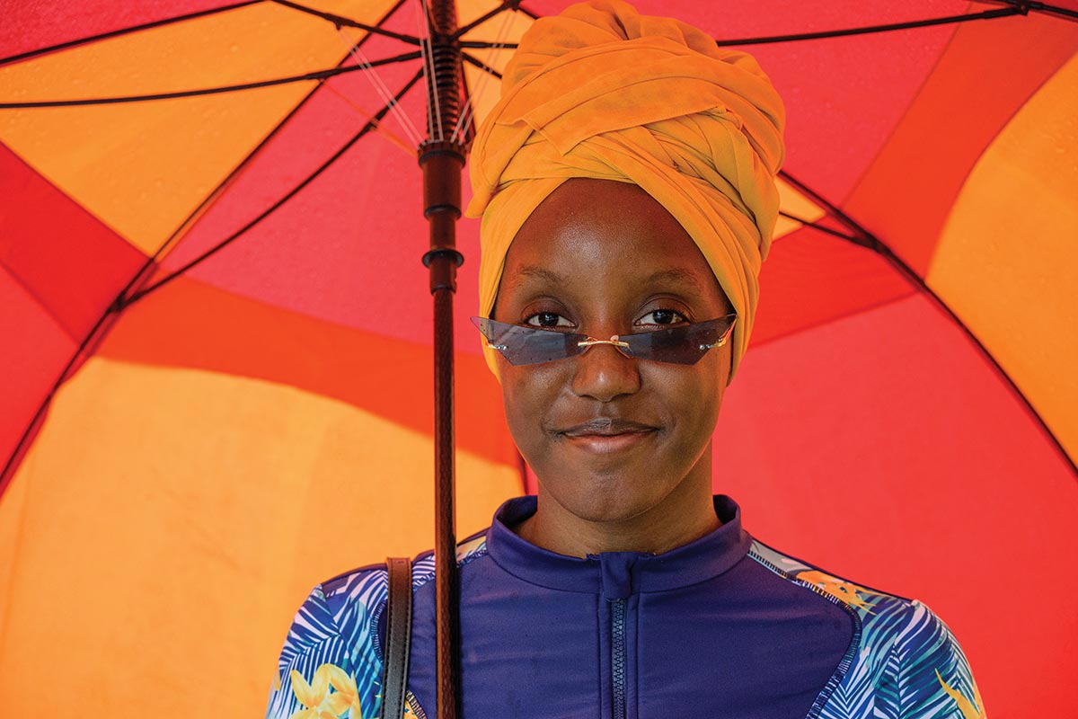 A Black woman with a yellow head wrap, blue top, and fashionable sunglasses stands under a vibrant yellow and orange umbrella that fills the background entirely.