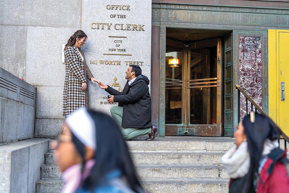 A man kneels as if proposing, holding the hand of his fiancée, at the top of the steps in front of the sign for the Office of the City Clerk of New York. Passers by are in the foreground.