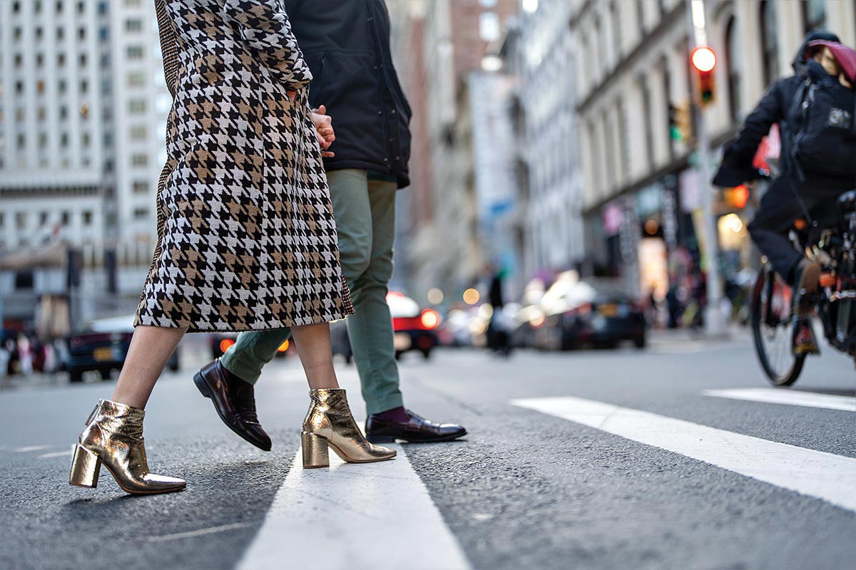 A couple walks on a city crosswalk, the photo focuses on their shoes, the woman's shiny gold short boots and the man's oxford wingtips.