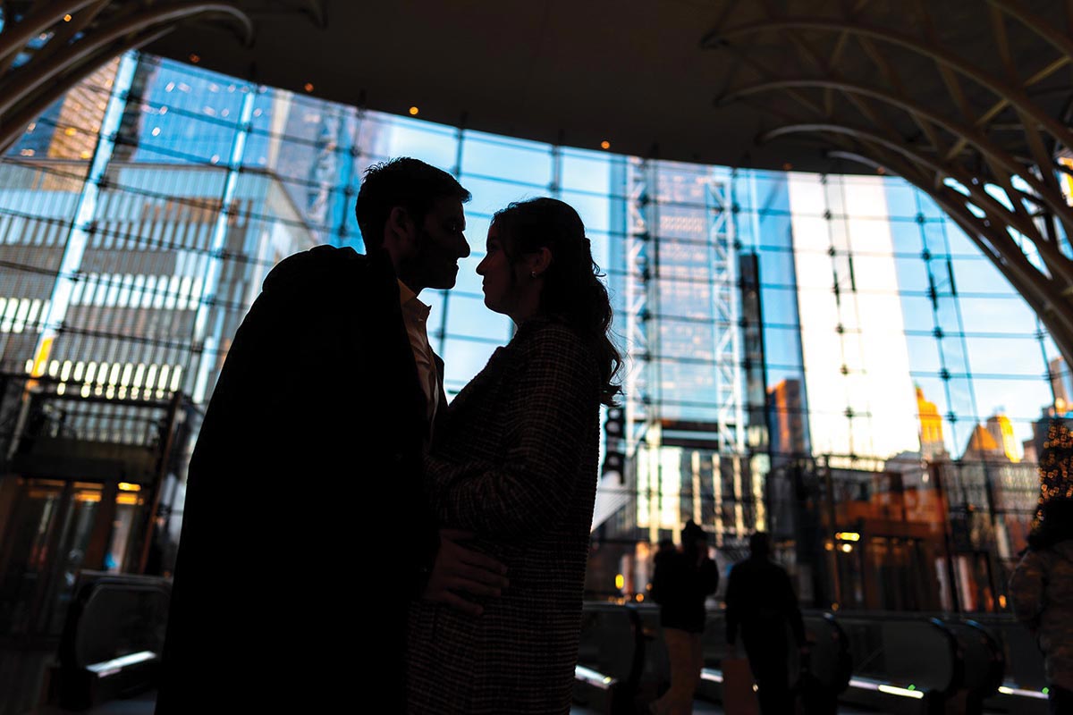 The silhouette of a couple stands out against the shiny backdrop of a glass building at sunset. A dark overhead structure frames the composition.