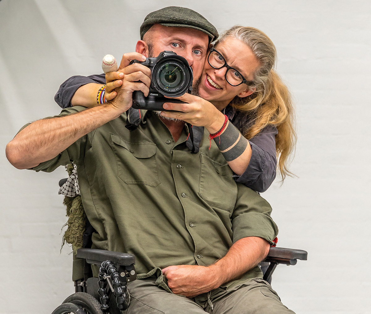 Uri Golman sits in a wheelchair and holds a camera, assisted by his wife Helle, who stands behind him.