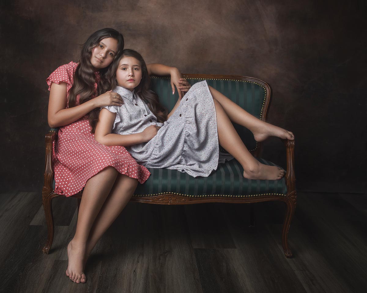 Two girls with long hair, wearing dresses, sitting on a loveseat