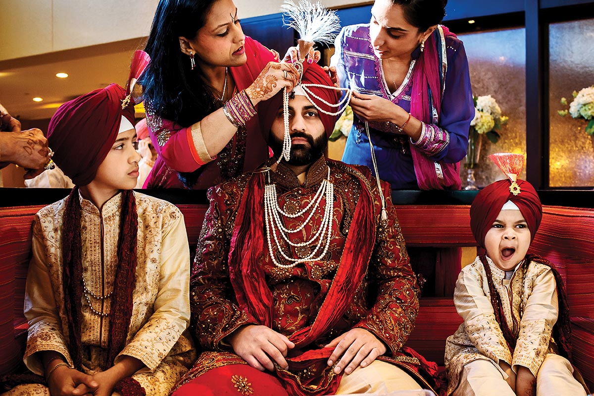 A family dressed in festive Indian wedding garb are centered around a man seated on a red couch. Two women standing behind the couch adjust ornaments on his turban. Two young boys flank him, seated on the couch. The boy on right yawns.