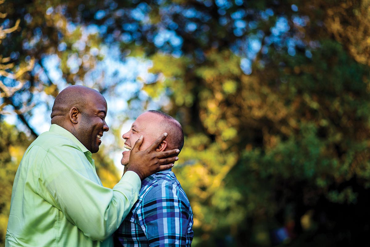 A male couple stand facing each other, profiles to the camera, smiling with joy in front of green trees that are out of focus in the background. 