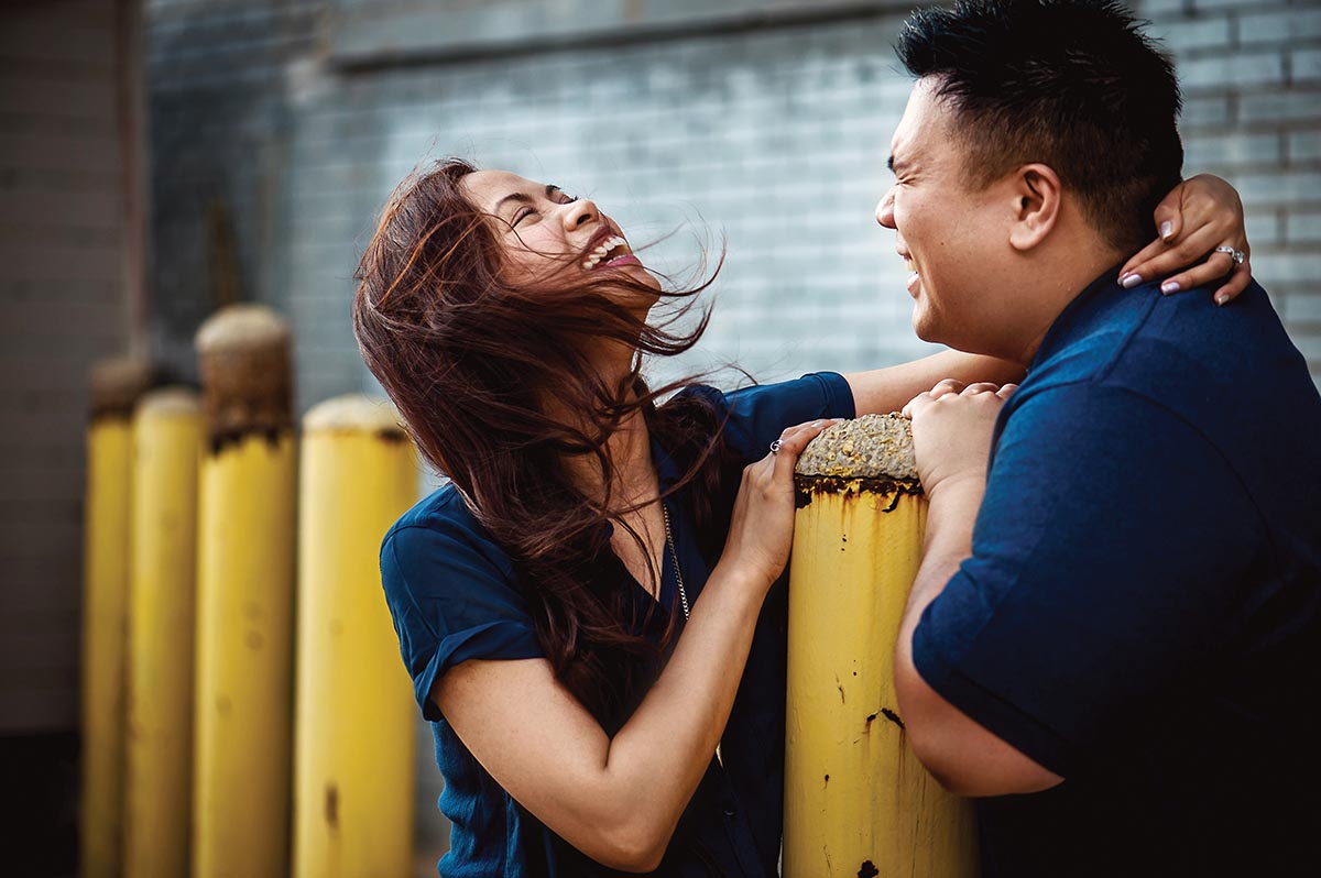 An Asian couple wearing blue shirts laugh together, standing on either side of a yellow concrete and metal loading dock pole barrier. Her left arm is draped over his right shoulder and behind his neck.