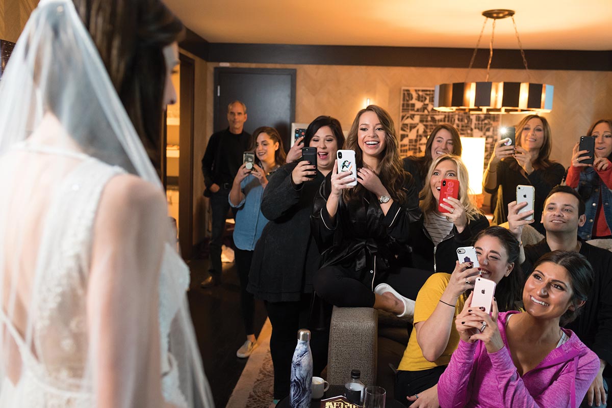 The bride's side of a wedding party is gathered in a hotel room, smiling and taking camera phone photos of the bride in the foreground, left.