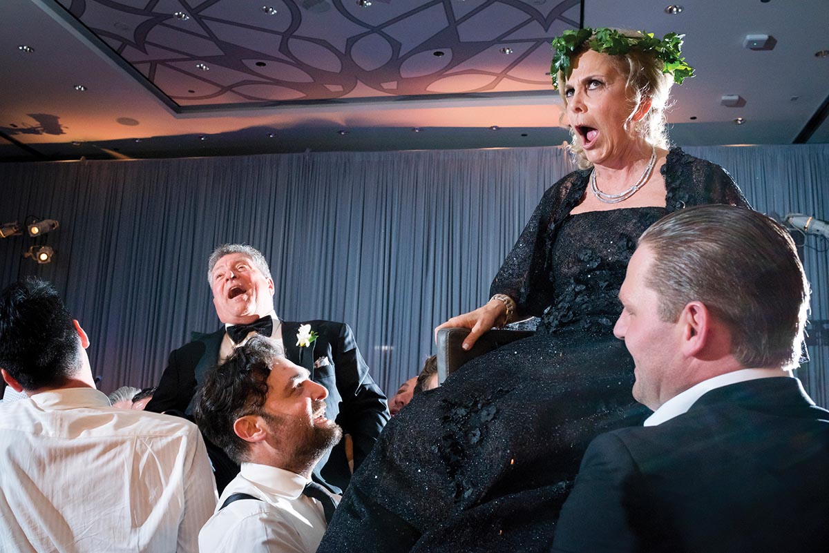 Parents in formal wear are lifted in chairs during the Hora dance at a wedding. The mother is making a funny face and the father is laughing.